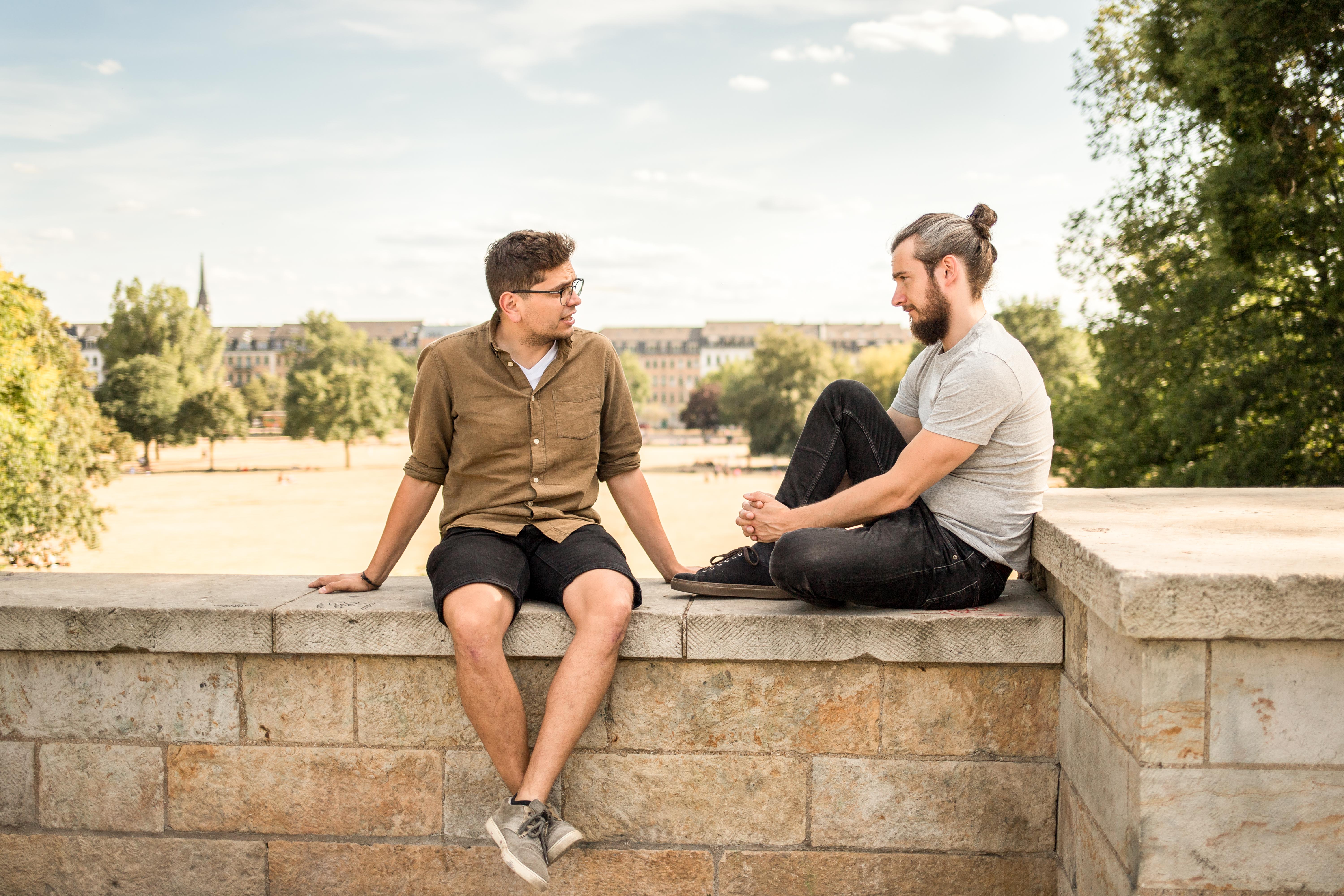 Foto von zwei jungen Männern, einander zugewandt auf Mauer sitzend, die sich unterhalten, im Hintergrund ist Park zu sehen