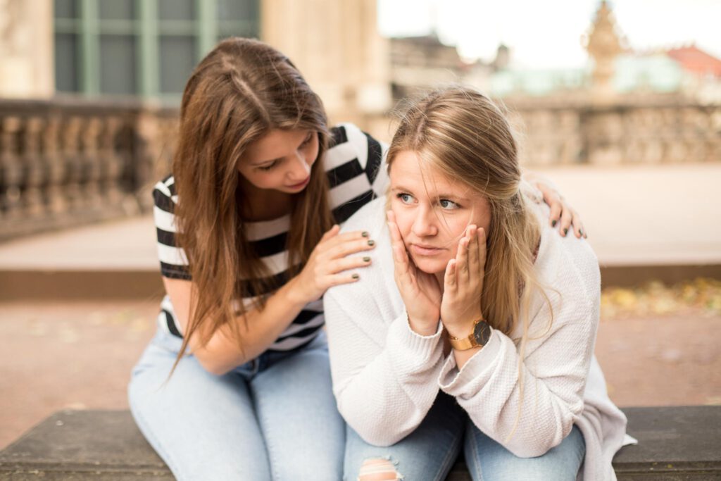 Foto von zwei jungen Frauen, nebeneinander sitzend, die eine ist nach vorne gebeugt und hat ihre Hände am Gesicht, die andere legt dieser Frau die Hände auf die Schultern