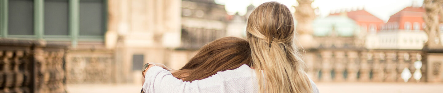 Foto von zwei jungen Frauen von hinten, eine Frau umarmt die andere, im Hintergrund sind Häusersilhouetten zu sehen